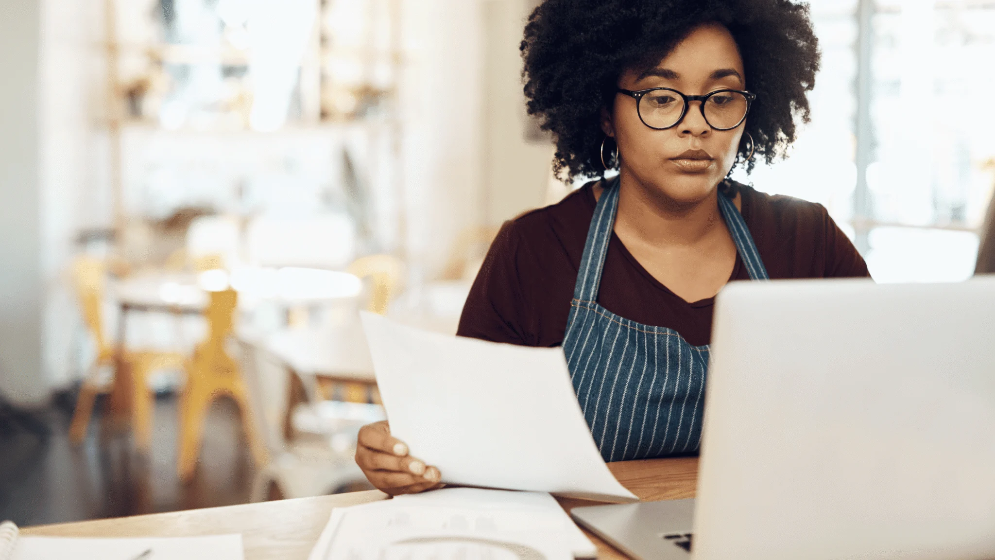 a-black-woman-with-an-apron-sat-processing-at-a-laptop-